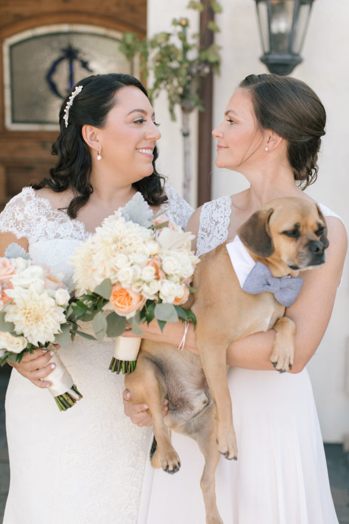 Two brides smiling at each other holding flowers and their small dog on their wedding day
