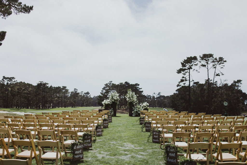 Wedding ceremony space set with large green and white foral arrangements on large tree trunks set on a golf course overlooking the ocean in California