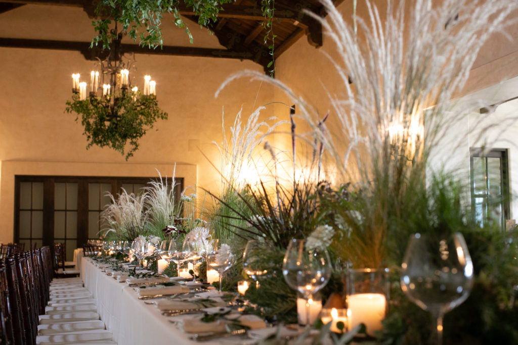 Large head table for wedding party with lush display of plants, grasses, florals, and candles with overhanging chandeliers with draped greenery