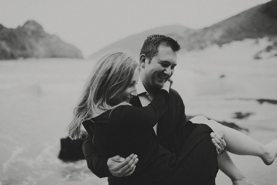 black and white engagement photo of couple on the beach where the groom is carrying the bride