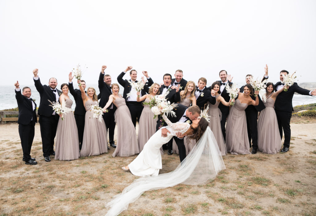 Wedding Party of bridesmaids and groomsmen celebrating as the bride and groom kiss dramatically in front of the ocean