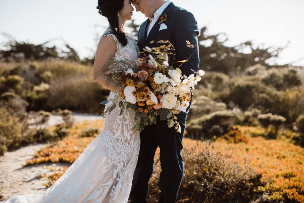 Bride and groom recently married embracing on sand with large boho style bouquet