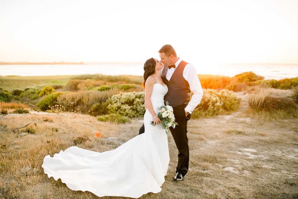 couple kissing at sunset in Spanish Bay pebble Beach. Bride is holding a bouquet of yellow and cream flowers.
