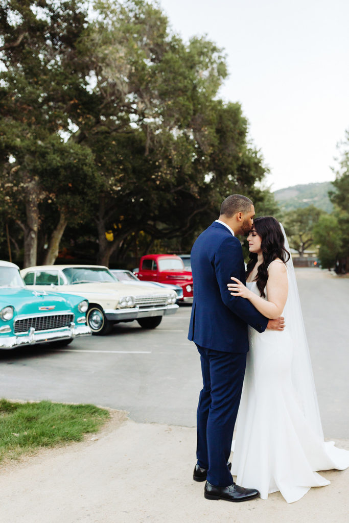 Bride and groom posing in front of their collection of vintage cars