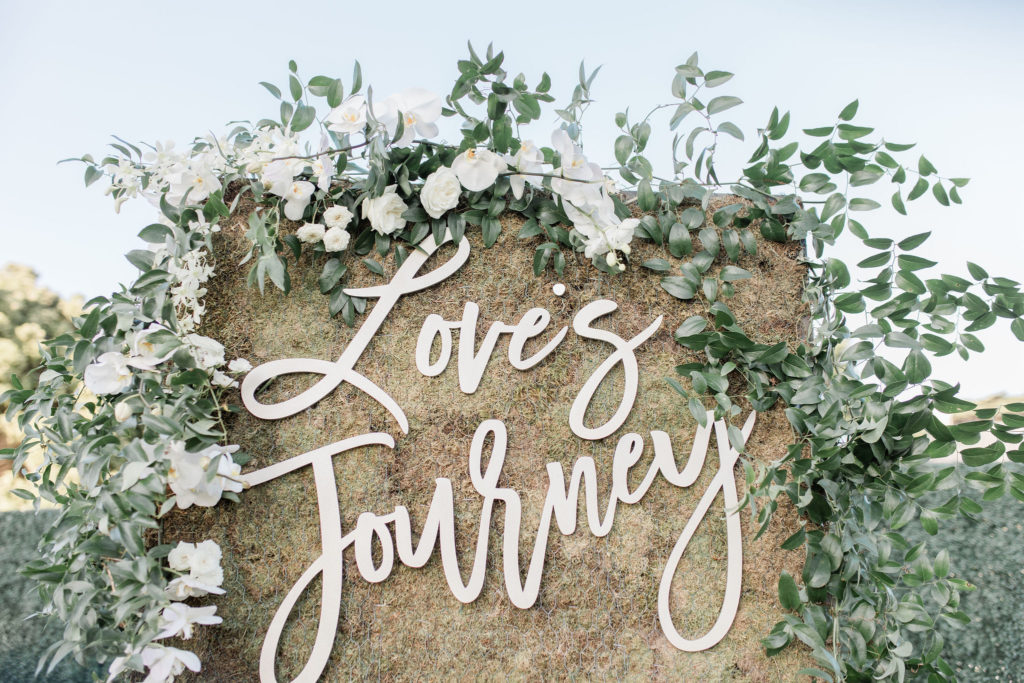 Escort card greenery wall with cursive text "Love's Journey"